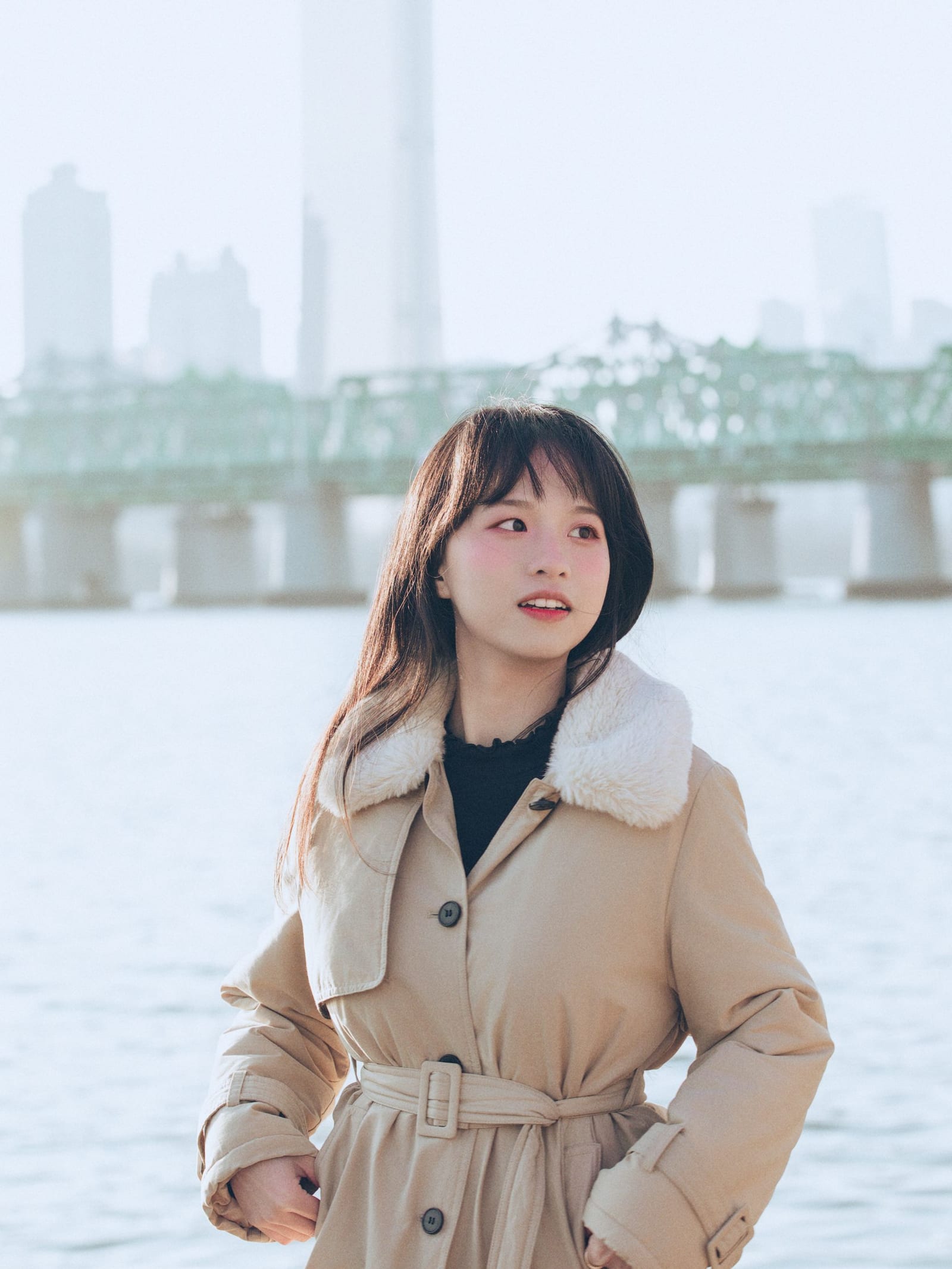 身着米色风衣的女性站在河边，远处是桥与城市天际线