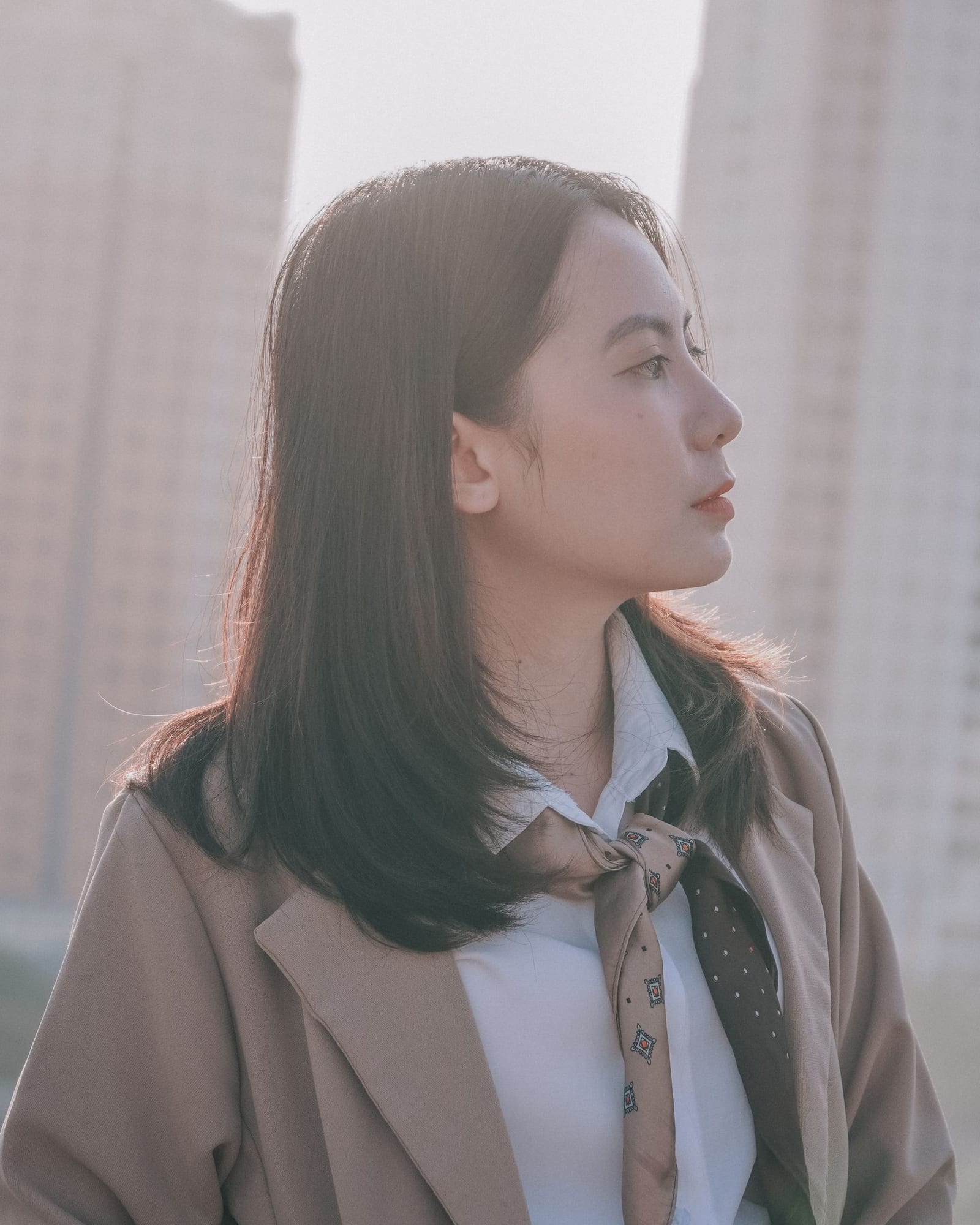 身着米色西装外套与丝巾的亚洲女性侧身立于城市楼宇之间，自然柔光